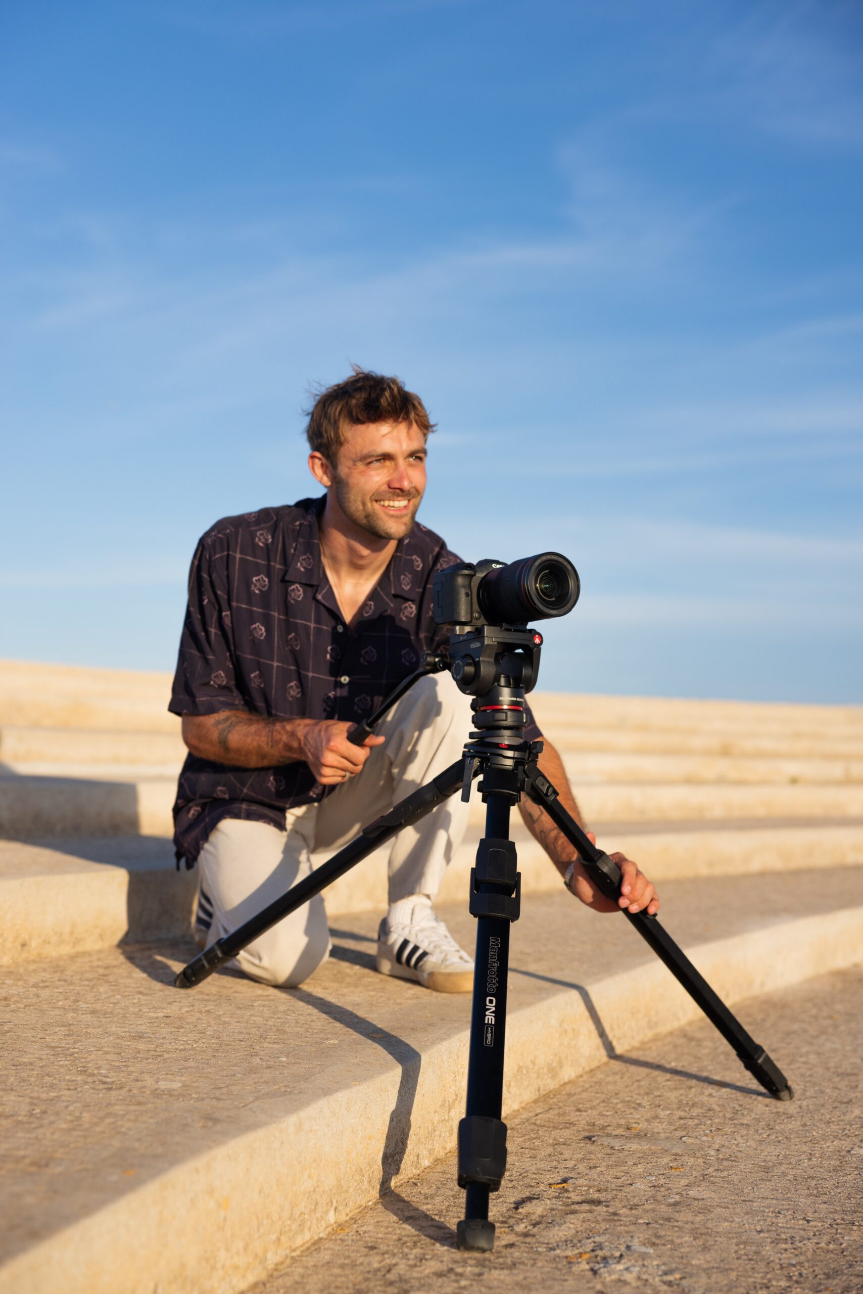 A man smiling and kneeling next to a camera on a mount, positioned on sandy steps
