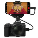 A small camera with a phone mounted to the top showing an image of a woman with oranges over her eyes