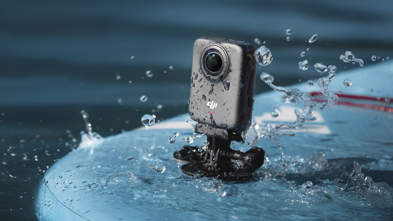 A small black DJI Osmo 360 camera stands on the end of a surfboard while water splashes it