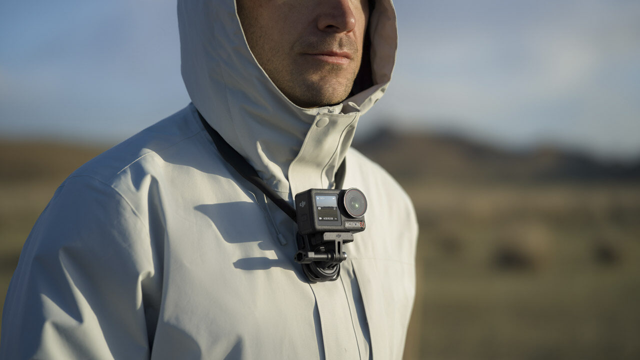 A man wearing a grey jacket stands outside with a small DJI Osmo 360 camera around his neck