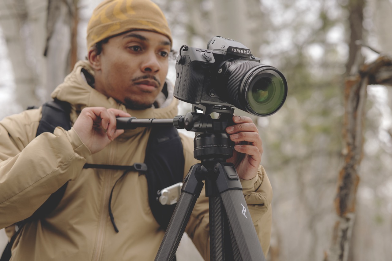 A man wearing a yellow beanie and coat stands outside, adjusting a camera on a tripod