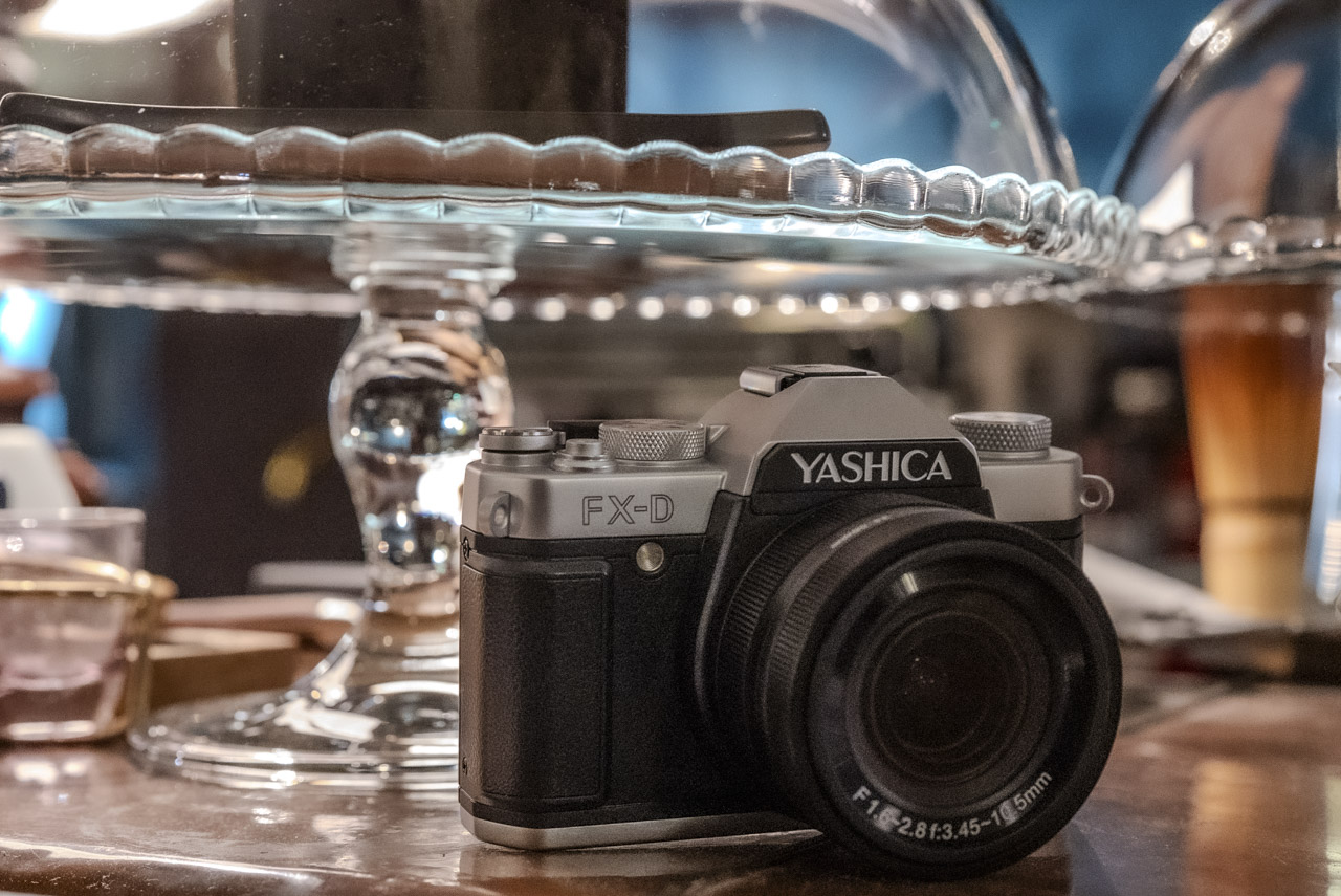 A black and silver Yashica camera sits on a wooden table in front of a glass cake stand