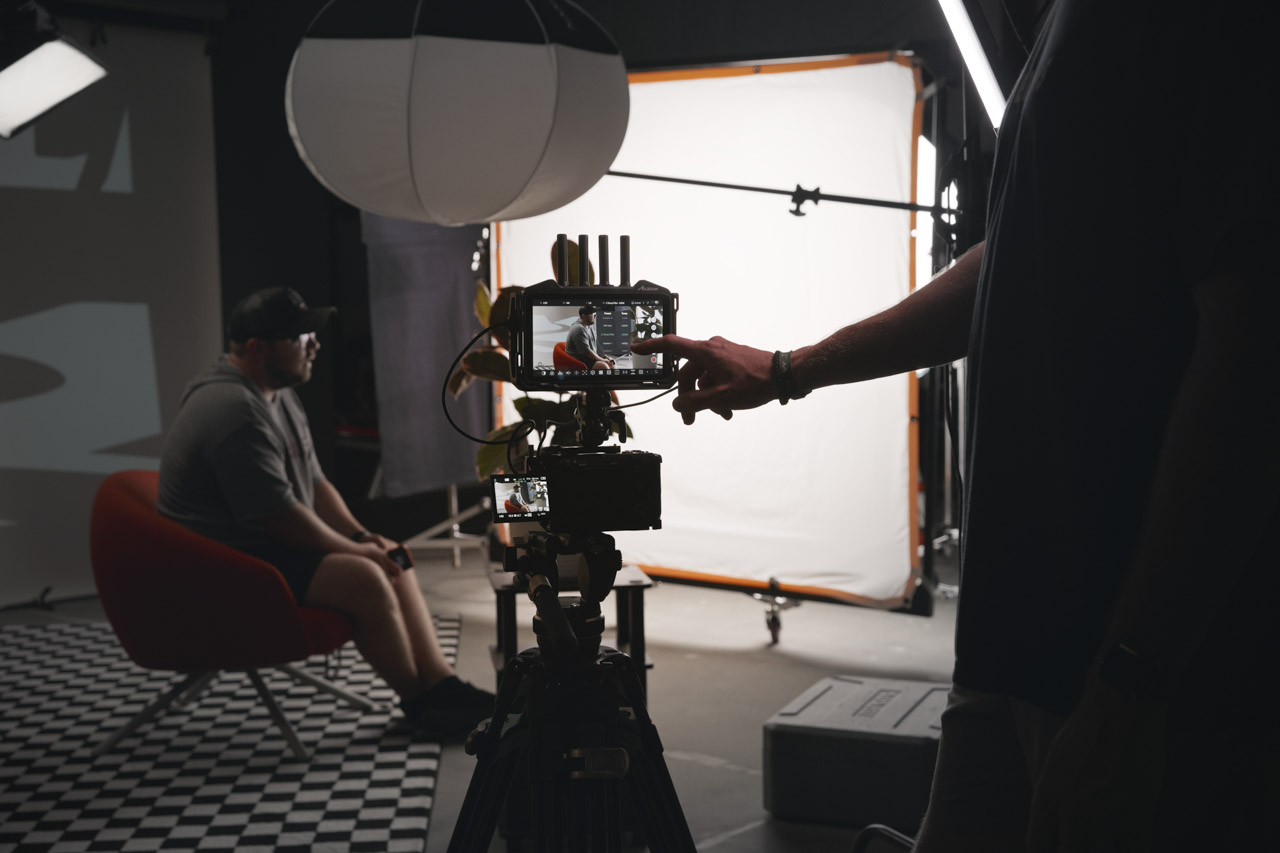 A camera with a touchscreen above. A person touches adjusts the touchscreen while a man sits on a seat being filmed