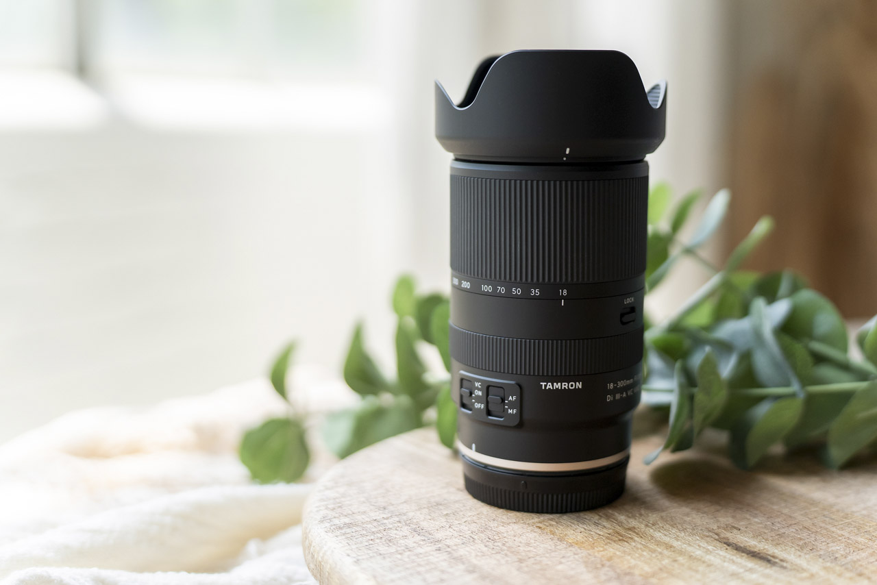 A black lens from Tamron sitting vertically on a wooden table with a branch behind
