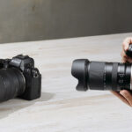A person holding a camera and large lens, taking a photo of another camera on a wooden table