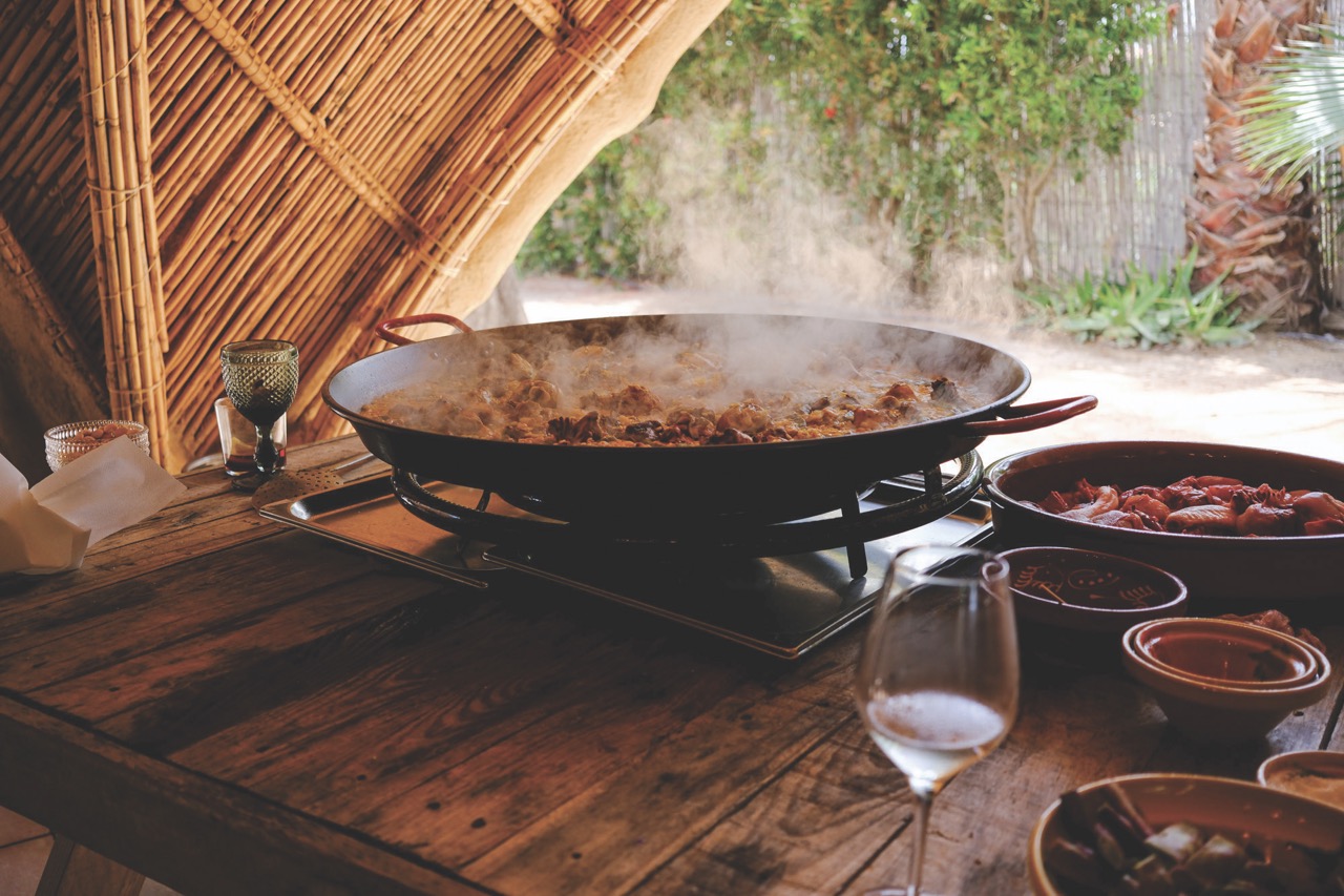 A steaming pot of food sits on a wooden table outside under a canopy