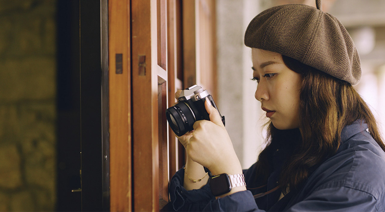 A woman wearing a beret stands in a doorway and looks closely at a small Yashica camera