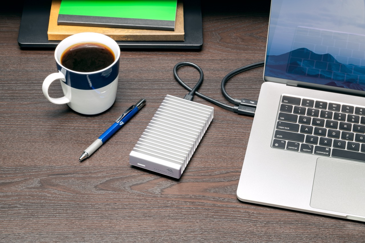 A white portable SSD from OWC attached to a laptop, next to a coffee and pen on a desk