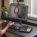 A man sits at a desk with a large monitor, keyboard and a colourful audio mixing board from Rode