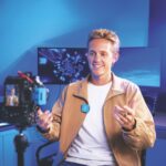 A smiling man wearing a brown jacket and blue Rode microphone, sitting in front of a camera in a blue room