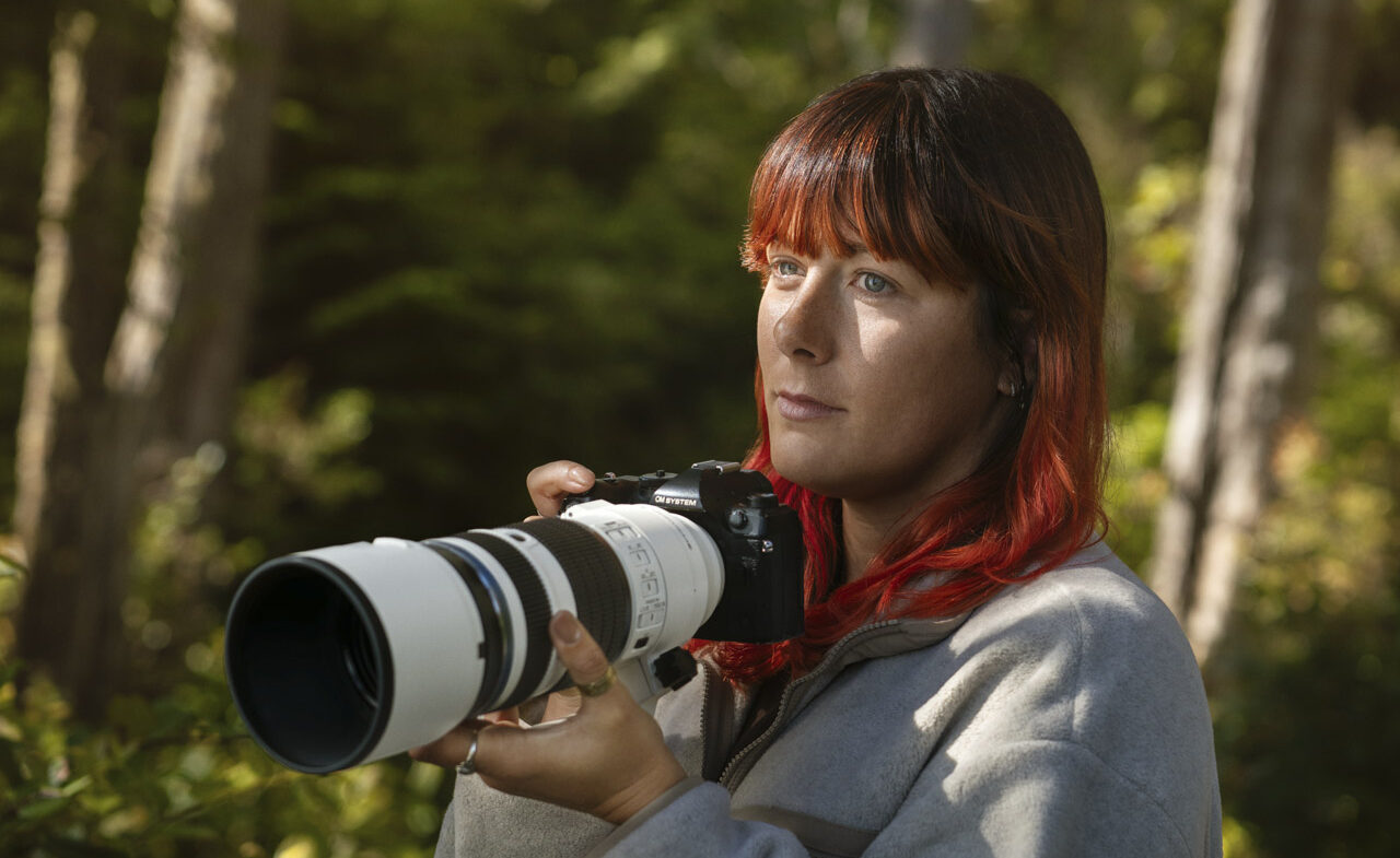 A woman with red hair holding a small camera with a large OM System telephoto lens attached. She stands in a forest