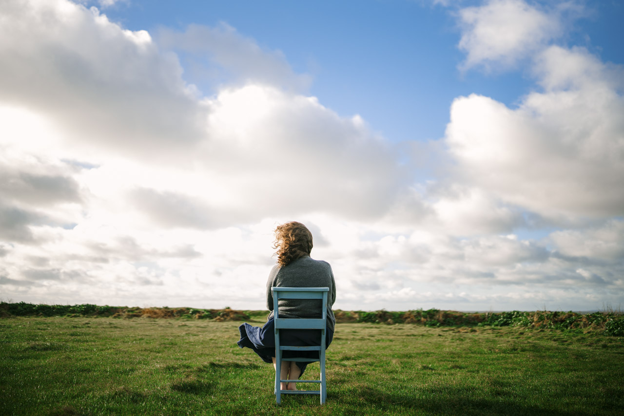 A person with long hair sitting on a metal chair in the middle of an empty field, under a cloudy sky