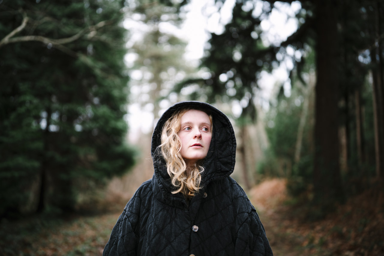 A blond girl wearing a large black hoodie. She stands between dense trees in a forest