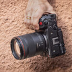 A hand holding a Canon camera, with an orange textured wall behind