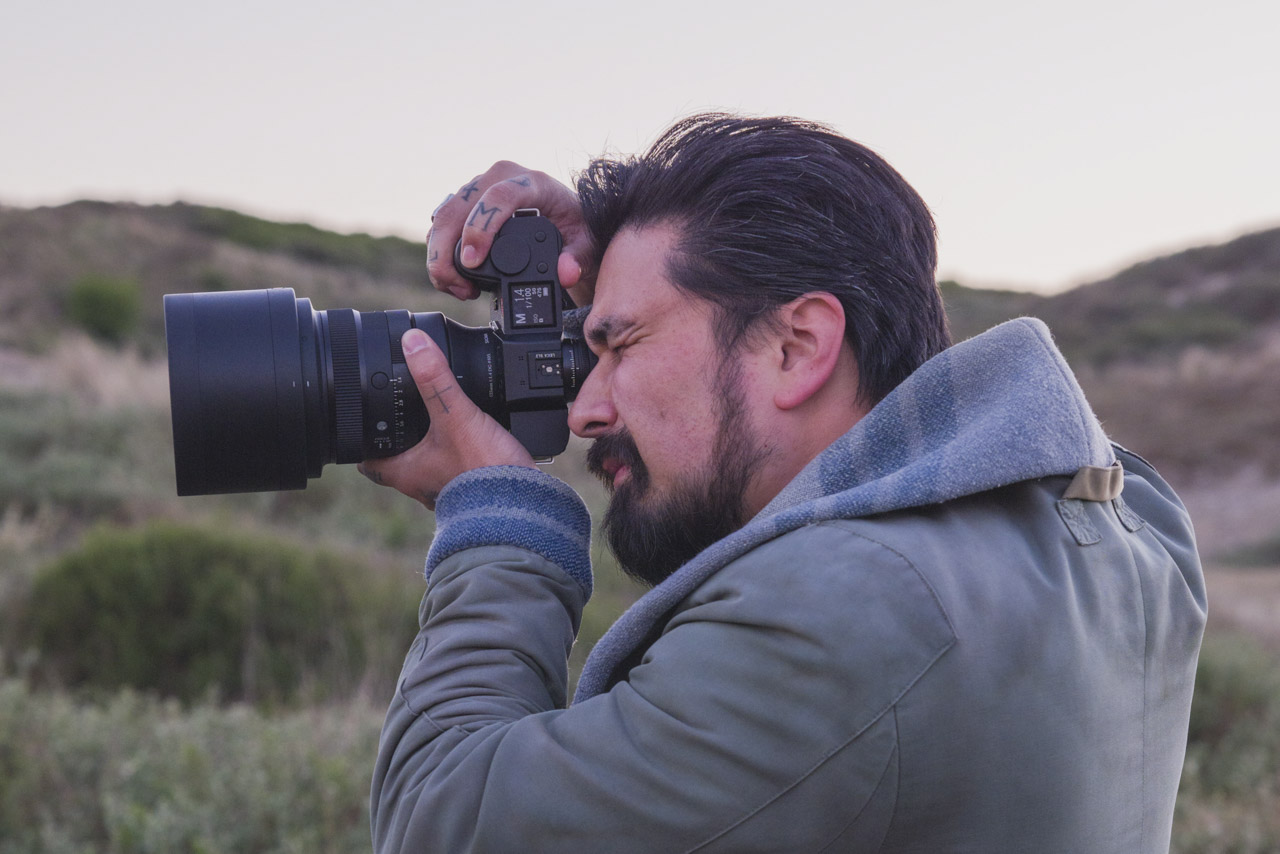 A man standing outside in the countryside and holding a camera up to his eye. The camera has a large prime from Sigma attached