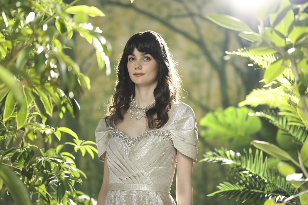 A woman wearing a satin dress standing in a gap between green trees, with sunlight streaming between the branches