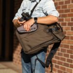 A man puts a camera in a brown bag from Think Tank Photo that is slung over his shoulder. The man stands in front of a brick wall