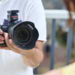 A person holding a Nikon camera and pointing it at a woman in yellow