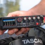 A hand holding a rectangle recording device from Tascam. The person is outside in a forest