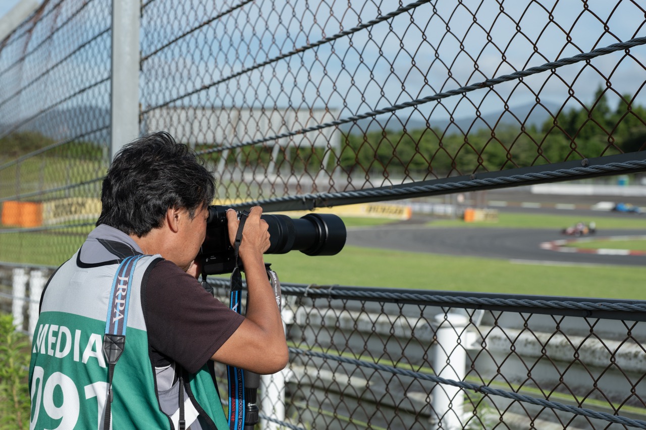 A man holding a camera with a large Panasonic lens attached up to his face. He wears a green bib and shoots a car racing track through a gap in a chainlink fence