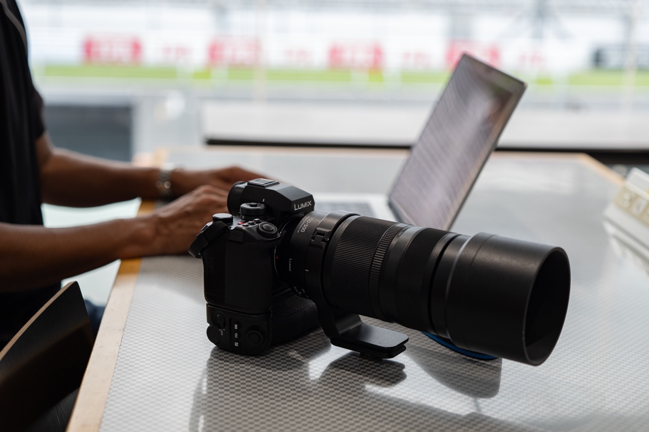 A camera with a Panasonic lens attached is sat on a shiny table, next to a person working on a laptop