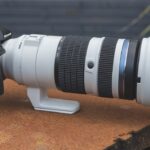 A black and white long lens from Om System, connected to a small camera and places on a wooden table