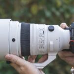 A professional white lens from Sigma attached to a small camera. It is being held up to a man's face