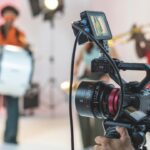 A Canon camera in focus with people setting up lighting equipment in the studio behind