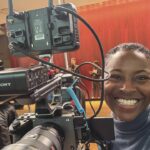 A young woman operating a camera and smiling in front of a stage