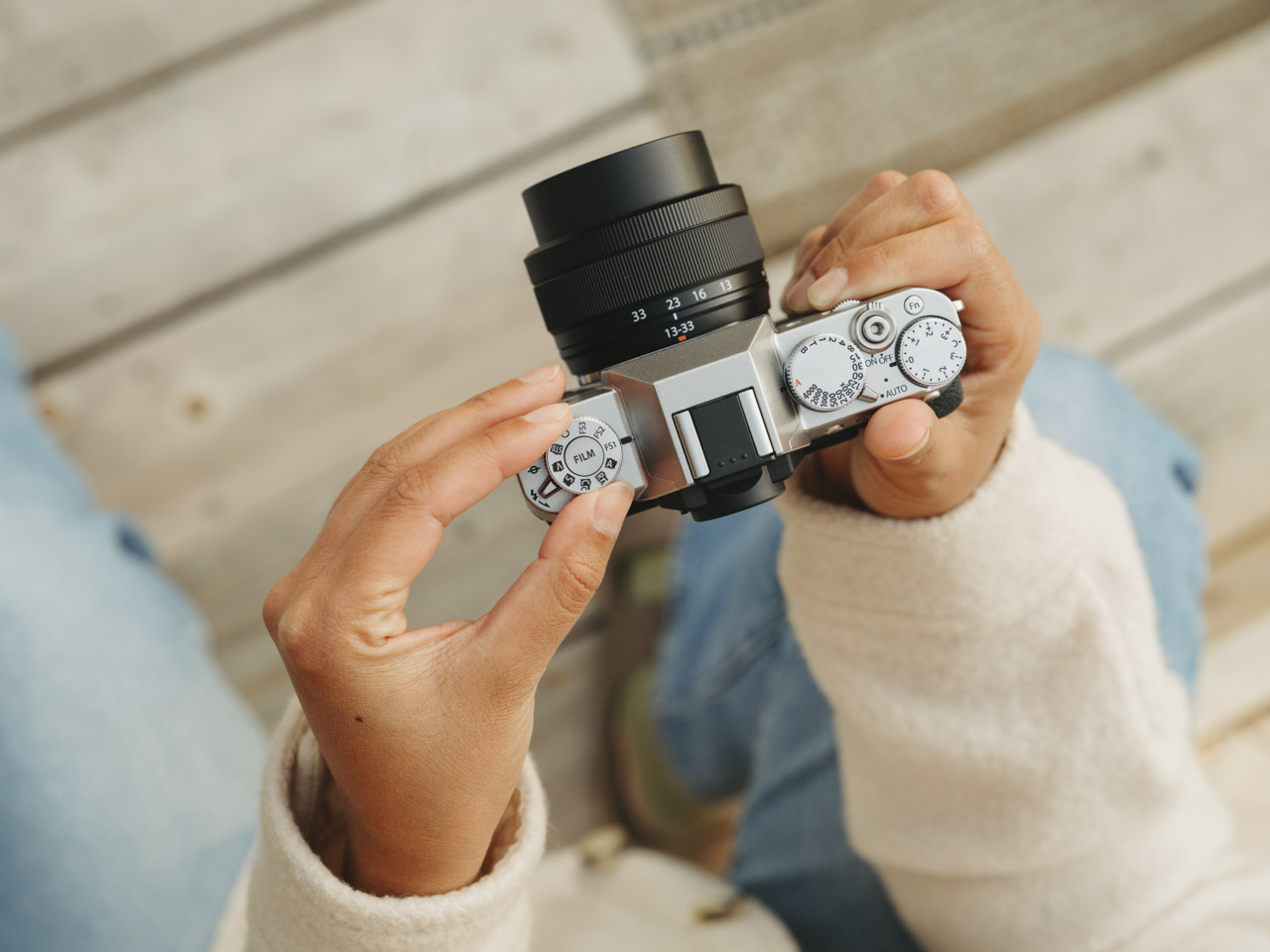 Top-view of a pair of hands holding a Fujifilm camera above wooden planks. The person wears blue jeans