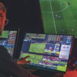 A person faces a computer screen and uses their mouse and a finger to adjust the videos of football on the screen. A larger screen in the background shows a football game