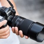 A person wearing white holds a Nikon camera with a Tamran lens attached. The camera is supported by a cord around the person's neck