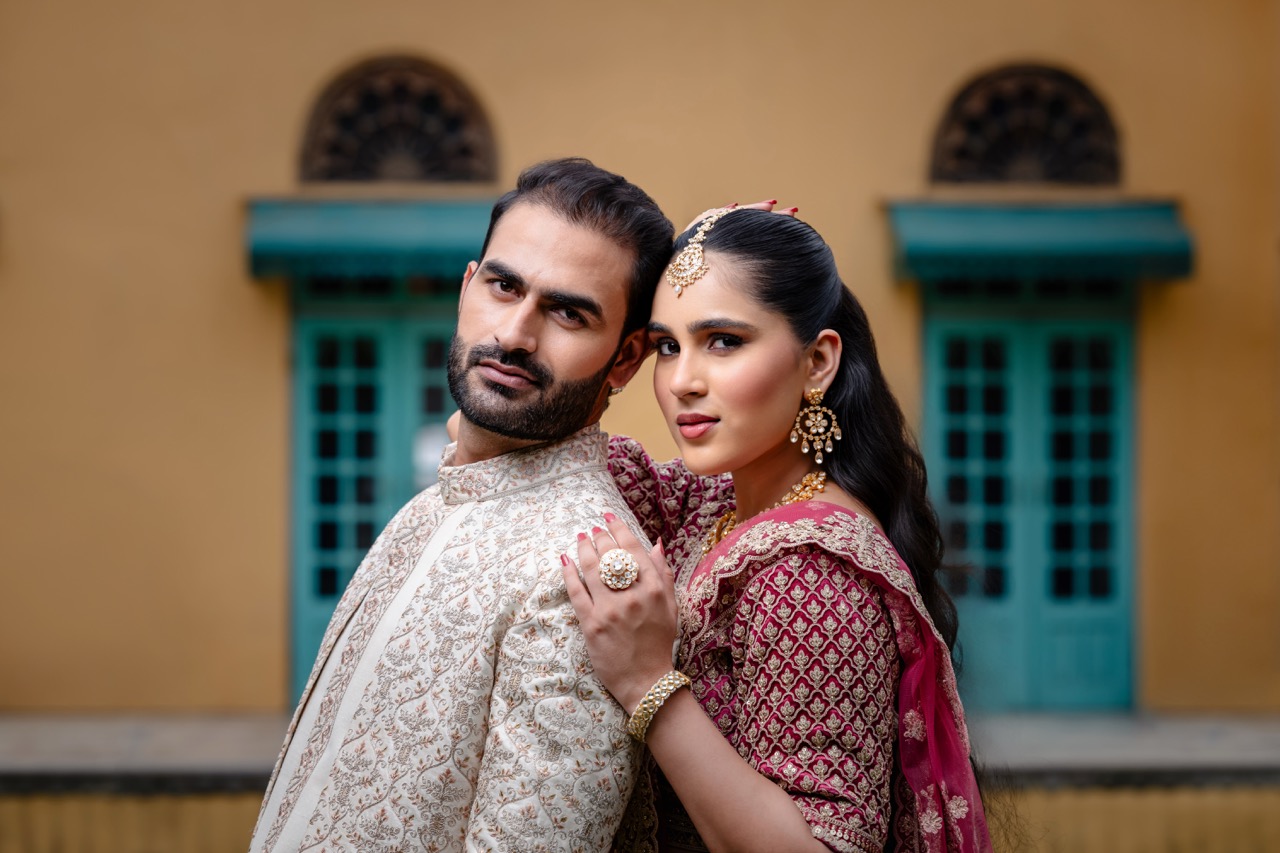 A woman holding a man's shoulders. They both wear wedding attire and stand outside a yellow building with blue windows