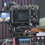 A professional camera with a Tascam recorder attached to the bottom. The camera films a blurred figure behind