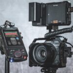 A camera with a device attached to the top stood next to a recorder from Tascam, against a grey background