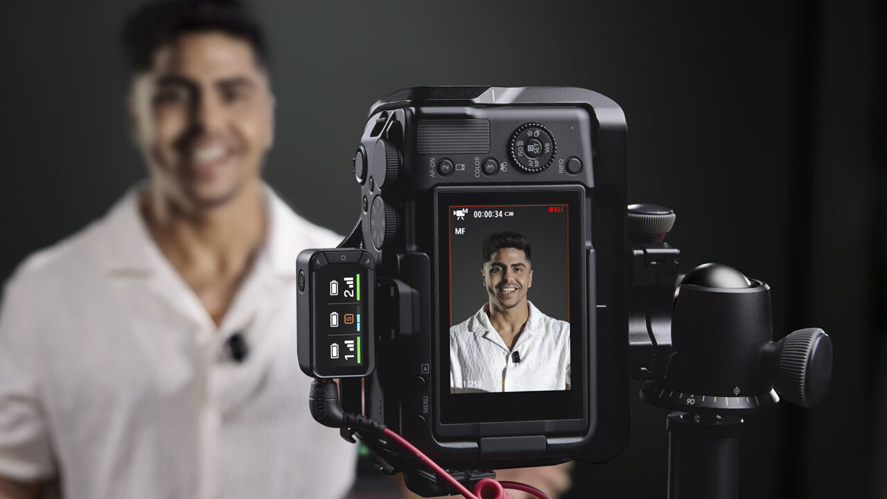 A camera with a Rode microphone attached to the side. The camera records a smiling man in a white shirt