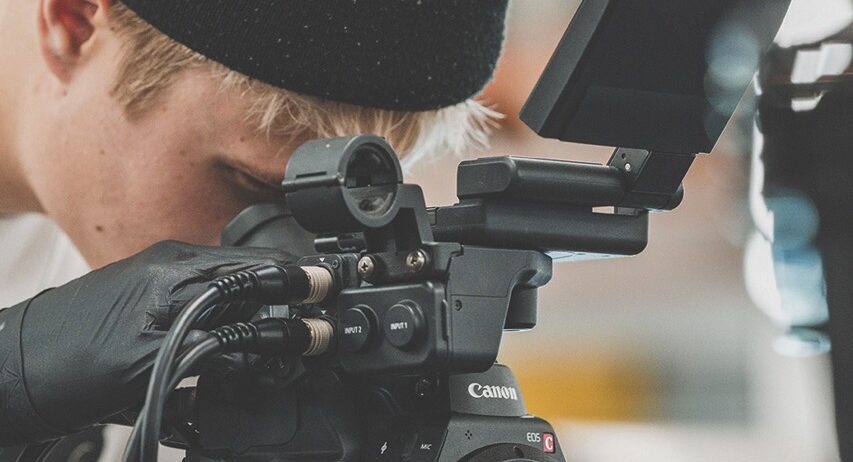 A man wearing a beanie and gloves looking through the viewfinder of a professional Canon camera