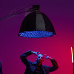 A man sitting on a chair in a red and blue studio with a large light from Harlowe overhead