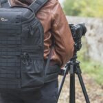 A person in a brown leather coat wearing black Lowepro backpack and operating a camera on a tripod outside