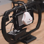 A white device hanging over the screen of a monitor in a holder, against a wooden desk