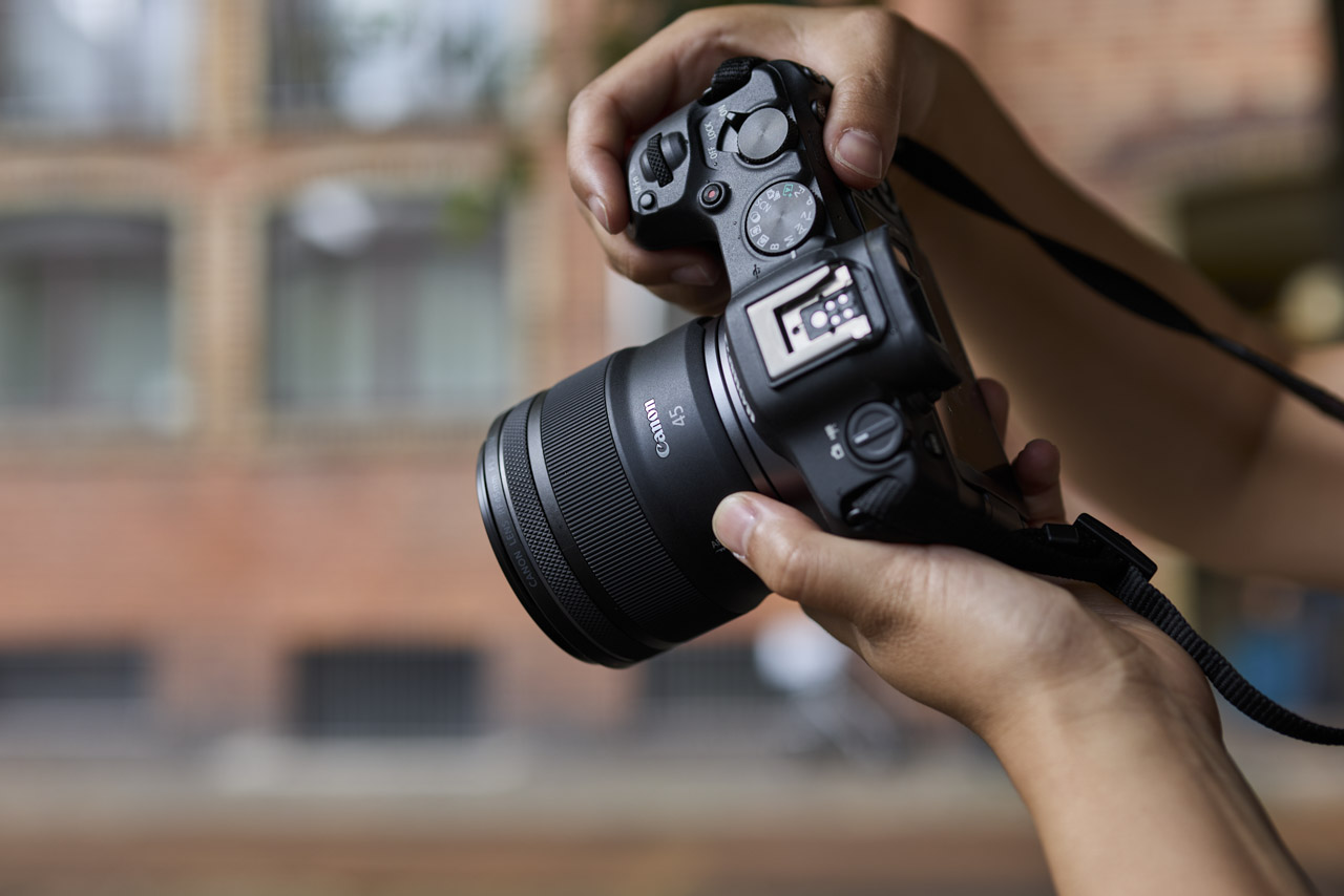A pair of hands holding a Canon camera and lens with a strap attached, against a blurred city landscape