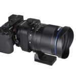 A black Laowa lens with a blue rim, balancing on a stand, against a white background