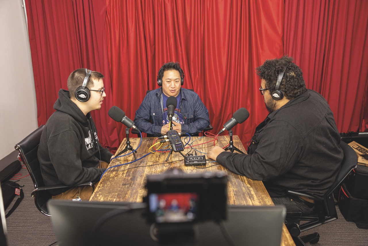 Three people sit around a wooden table with microphones and headphones, with a red curtain behind them