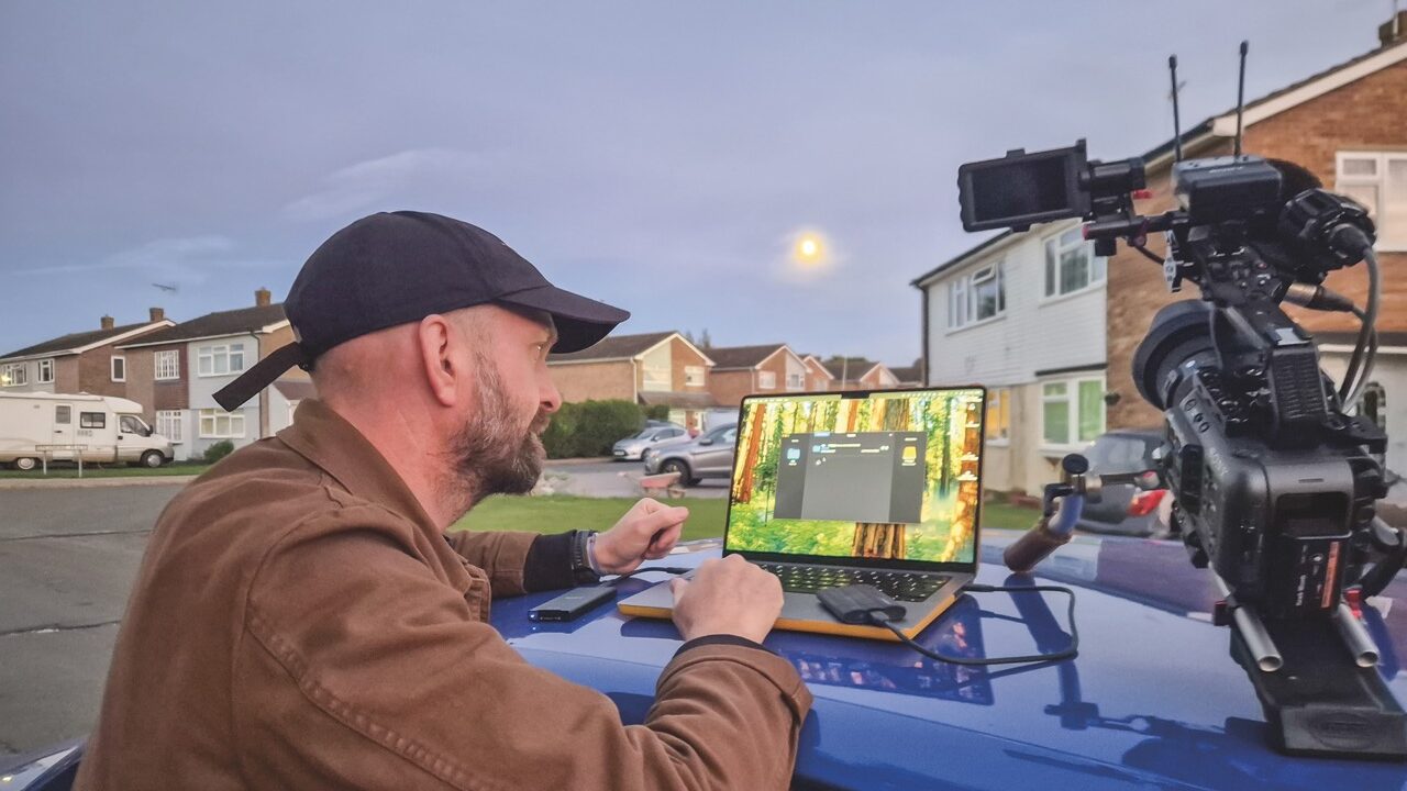 A man in a black cap operating a laptop, a professional camera and storage device on the top of a car