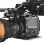 A professional camera from Blackmagic Design with an orange lens and a screen, against a white background