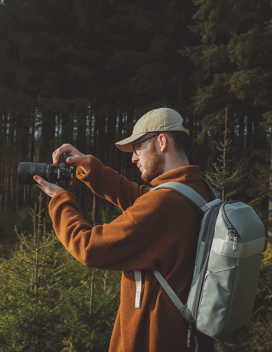 Side view of a person wearing a cap and backpack, holding a camera in a forest setting