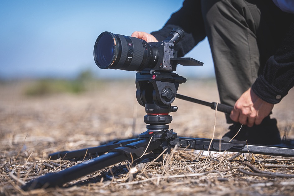 A person adjusting a camera on a low tripod, placed on brown grass