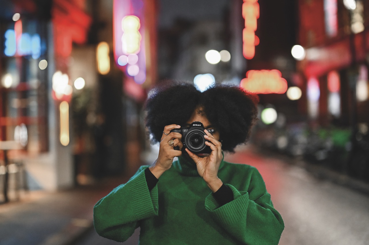 A person in a green jumper holding up a Nikon camera to cover their face, with a blurred city scene behind them