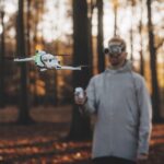 A person standing in a forest at dusk while wearing a headset and operating a drone