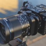 A black lens from Laowa attached to a camera and a tripod, against a blurred background